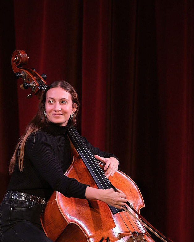 A female cellist, wearing a black top and black trousers, looking beyond the stage smiling, with a red velvet curtain behind her